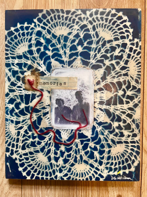A square mixed-media collage artwork featuring a cream-colored lace doily pattern over a dark navy blue background, with a central clear photo slot holding a vintage black-and-white portrait, a small paper tag reading 'memories,' and a red cord running through the center. The piece is photographed flat on a light wood surface.
